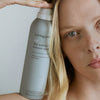 Nahaufnahme einer Frau, die eine Spraydose „Living Proof Full Dry Volume & Texture“ neben ihrem Gesicht hält.