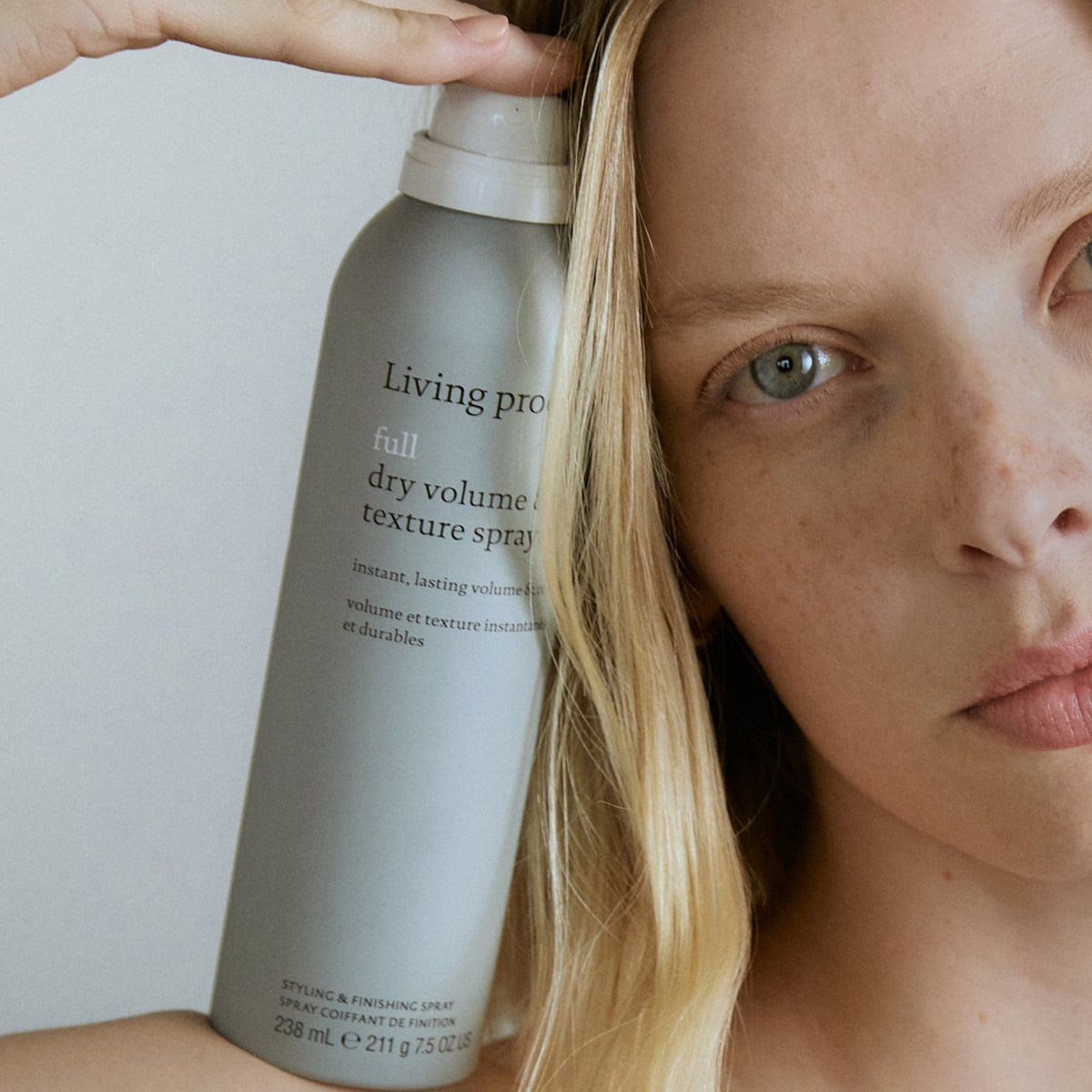 Nahaufnahme einer Frau, die eine Spraydose „Living Proof Full Dry Volume & Texture“ neben ihrem Gesicht hält.