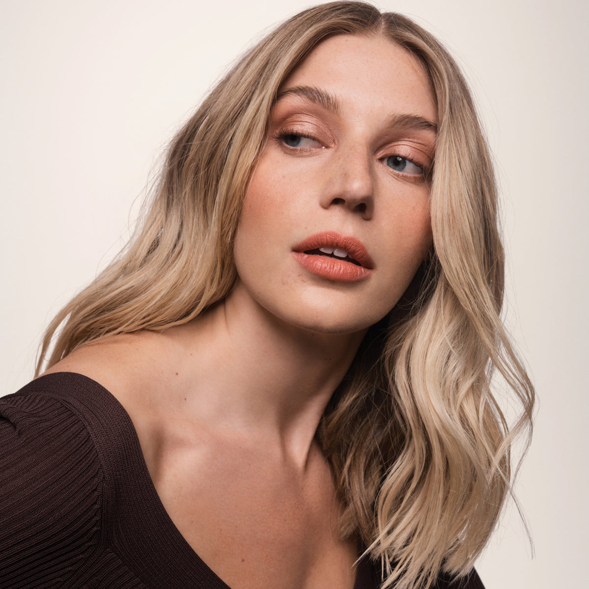 Eine Frau mit welligem, blondem Haar, das mit Living Proof Full Texturizing Foam gestylt wurde, blickt vor einem schlichten, hellen Hintergrund zur Seite.