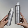 Zwei Hände halten silberne Flaschen Living Proof Perfect Hair Day Advanced Clean Dry Shampoo vor einem neutralen Hintergrund.