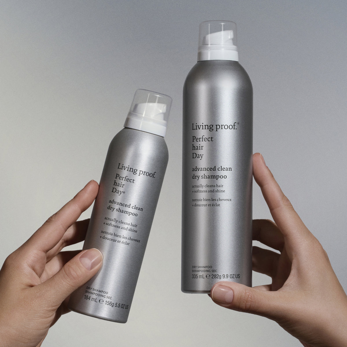Zwei Hände halten silberne Flaschen Living Proof Perfect Hair Day Advanced Clean Dry Shampoo vor einem neutralen Hintergrund.