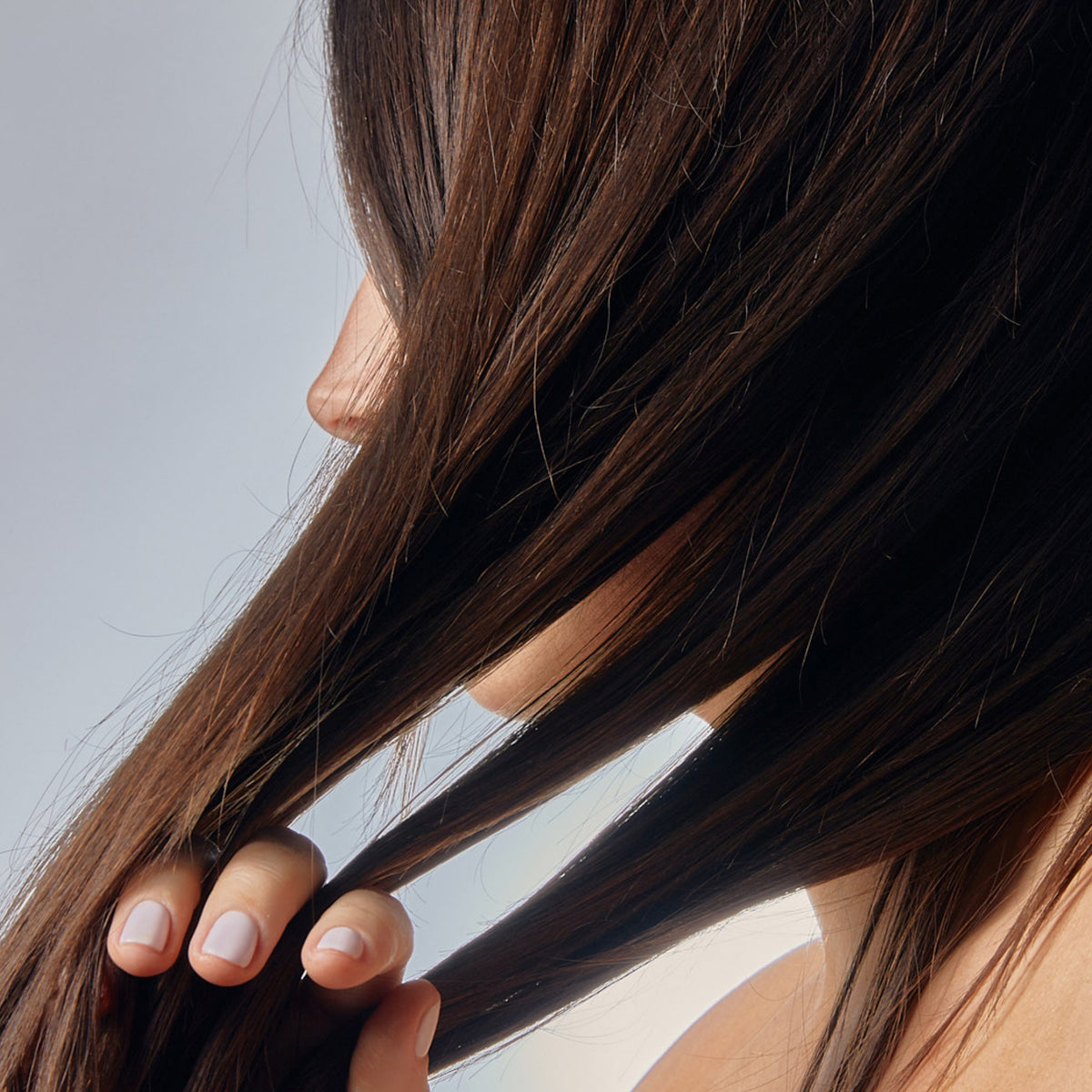 Nahaufnahme einer Frau mit langen braunen Haaren, die Strähnen in der Hand hält und die Wirkung der Living Proof Restore Repair Mask demonstriert.