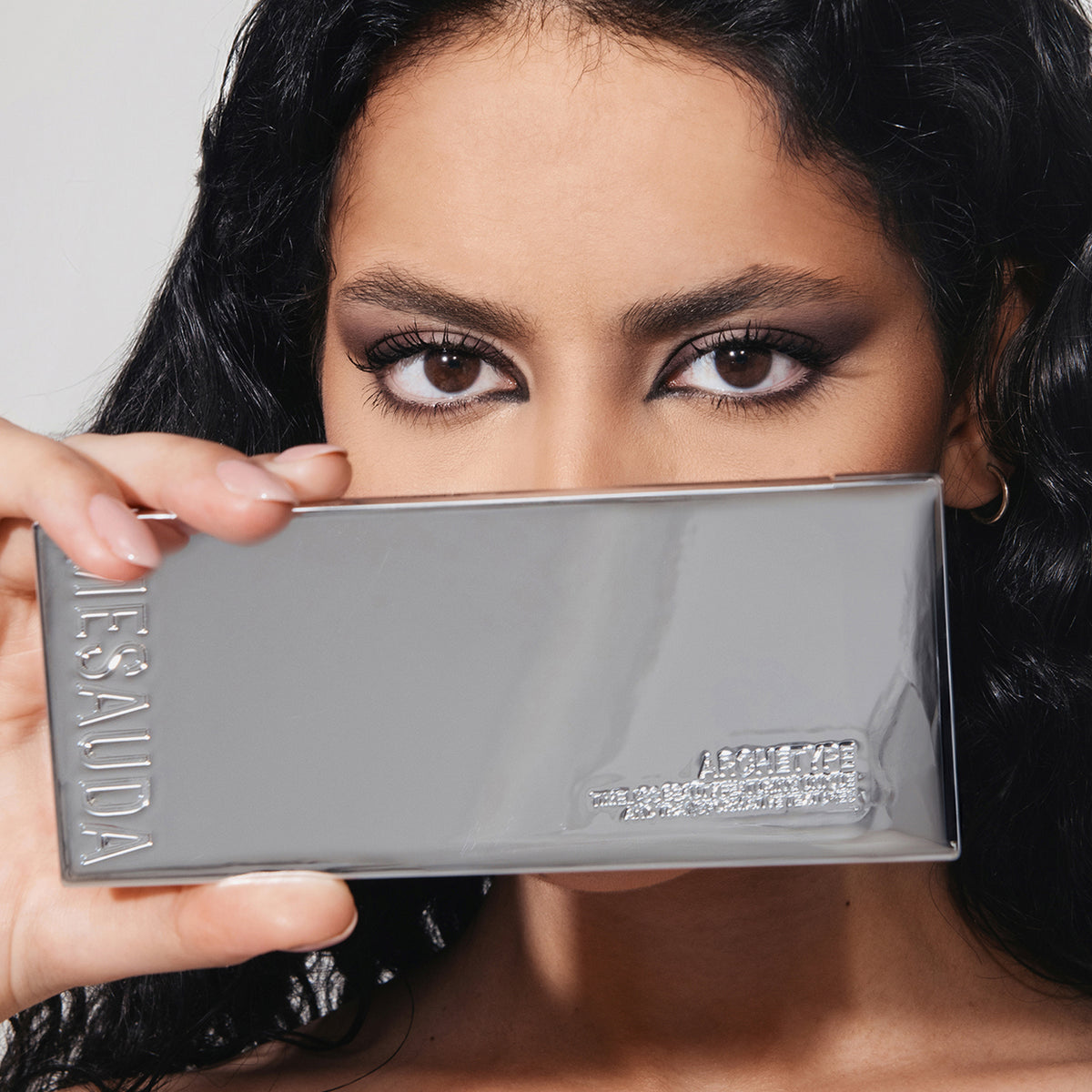 Eine Frau mit kräftigem Augen-Make-up hält sich die reflektierende silberne ARCHETYPE-Palette von Mesauda Beauty vor das Gesicht.