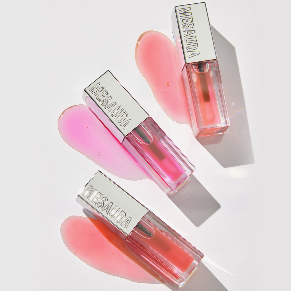 Drei Mesauda Beauty LIPOILOGY™ Lipgloss-Tuben mit verschüttetem rosa und pfirsichfarbenem Gloss auf einer weißen Oberfläche und weiche Schatten.