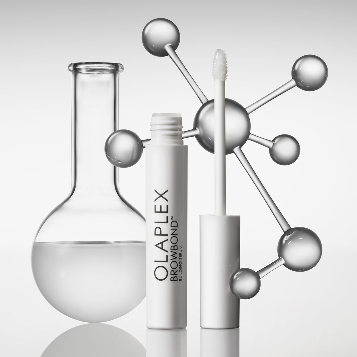 Eine weiße Browbond Building Serum™-Tube mit Applikator steht neben einem Glaskolben und einem metallischen Molekülmodell.