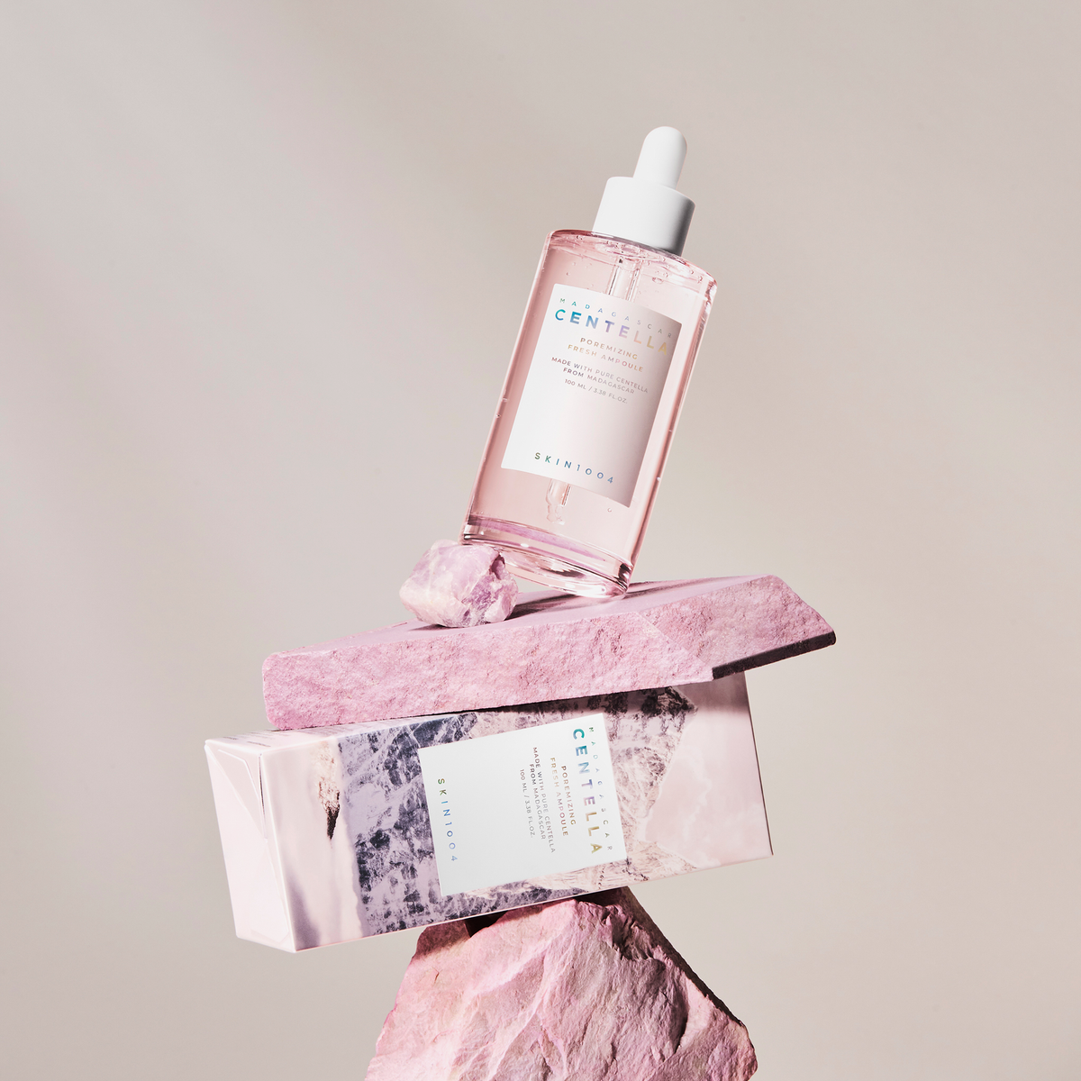 Die Madagascar Centella Poremizing Fresh Ampoule in einer rosafarbenen Flasche mit Schachtel wird auf gestapelten rosa Steinen vor einem hellen Hintergrund präsentiert.