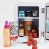 Beauty Fridge, geschmückt mit Hautpflegeprodukten, einer White Claw-Dose, einem Starbucks-Becher und Erdbeeren auf einer weißen Oberfläche.