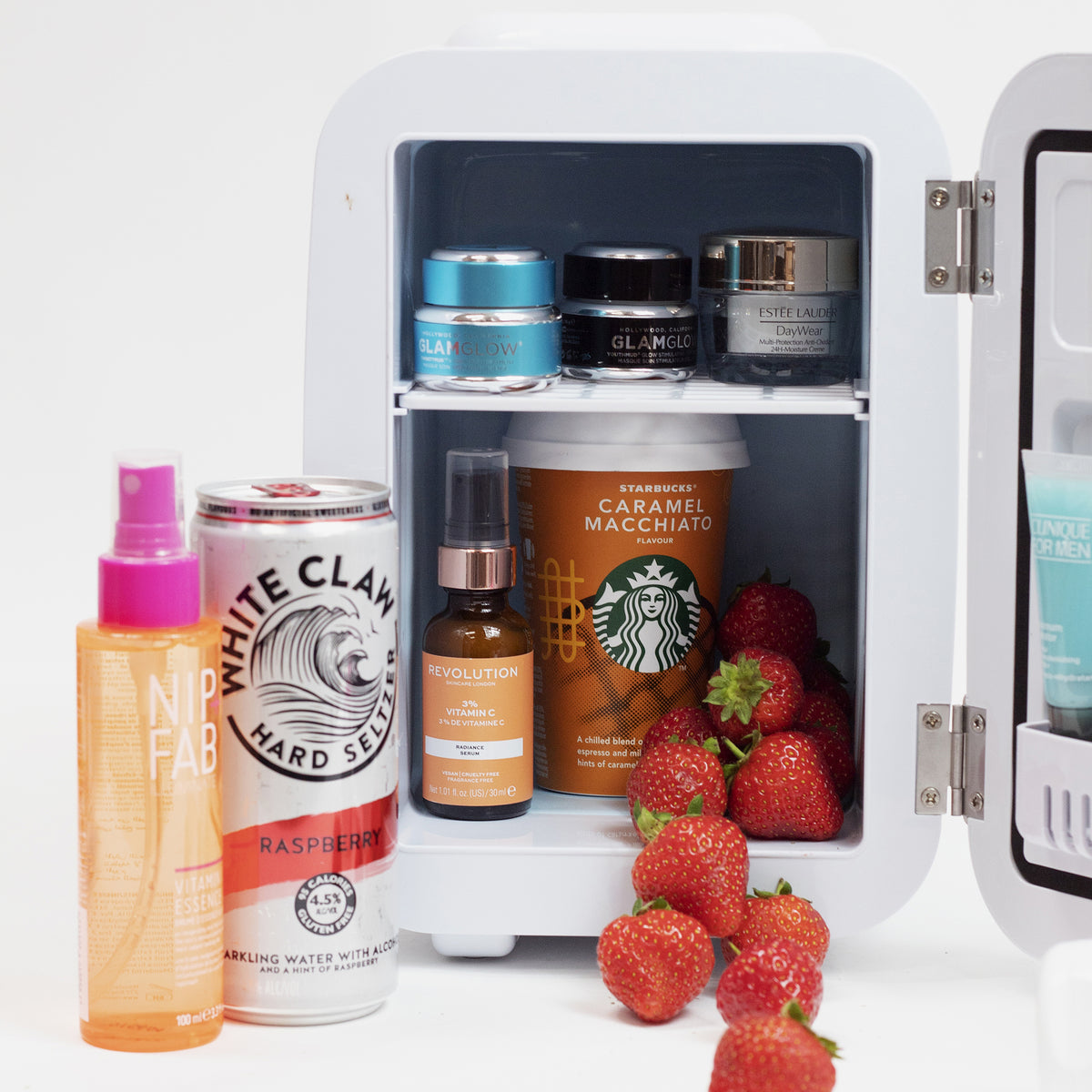 Beauty Fridge, geschmückt mit Hautpflegeprodukten, einer White Claw-Dose, einem Starbucks-Becher und Erdbeeren auf einer weißen Oberfläche.