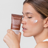 Mit geschlossenen Augen hält eine Frau eine Tube Bakuchiol Firming Cream in der Hand und streicht ein wenig davon über ihre Wange.