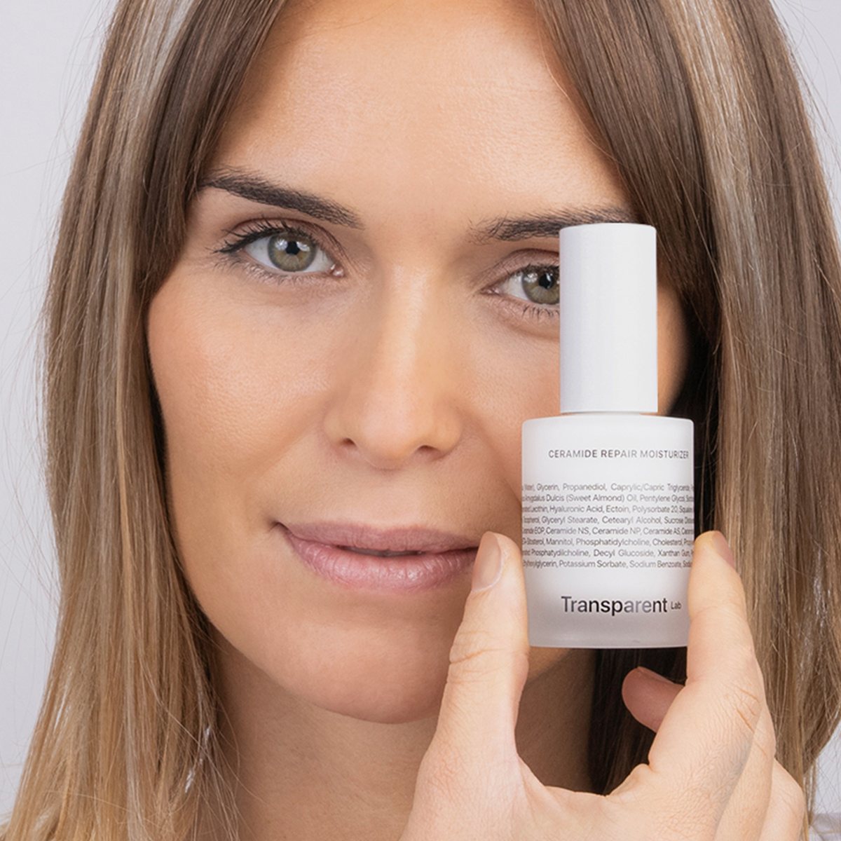 Eine Frau hält eine Flasche Ceramide Repair Moisturizer vor ihr Gesicht und blickt in die Kamera.