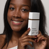 Eine lächelnde Frau hält die getönte Sonnencreme „Transparent Lab Mineral Tinted Sunscreen“ dicht an ihr Gesicht, vor einem schlichten Hintergrund.