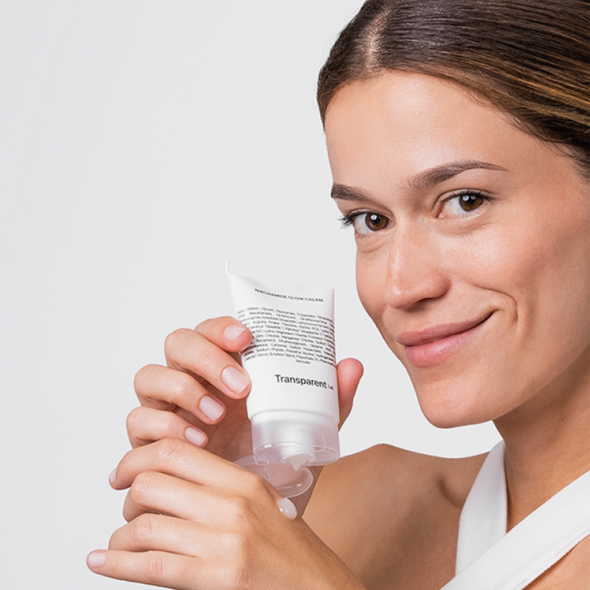 Lächelnde Frau, die die Flasche Niacinamide Glow Cream mit dem Produkt auf den Fingern vor einem weißen Hintergrund hält.