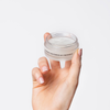 Eine Hand hält einen offenen Tiegel Overnight Restoring Lip-Treatment vir grau-weißem Hintergrund.