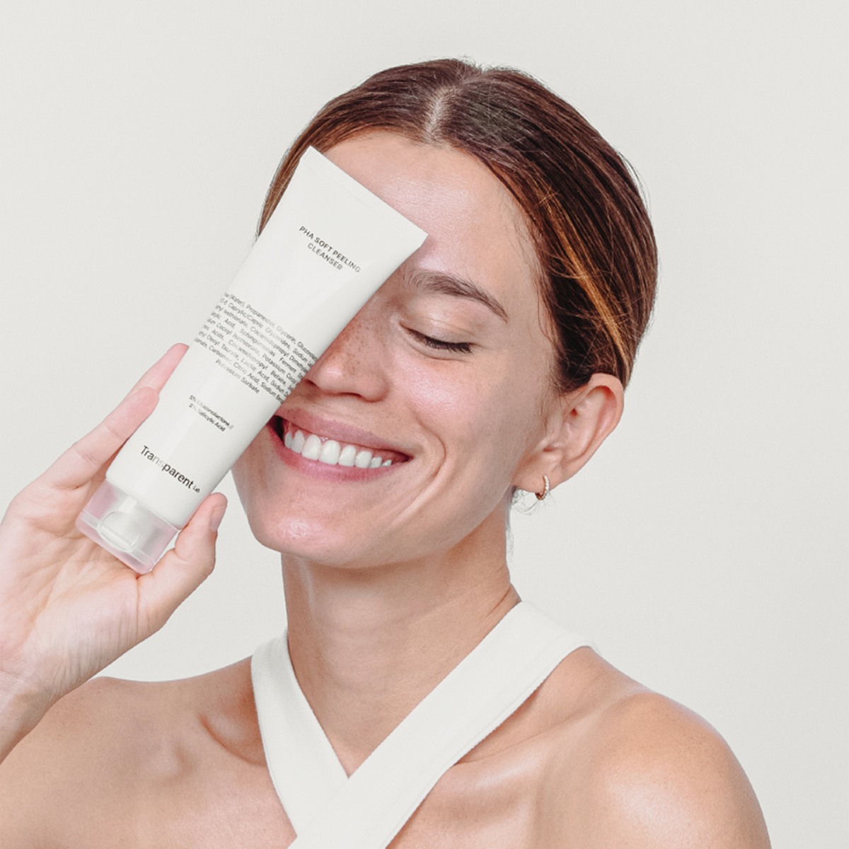 Eine lächelnde Frau in einem weißen Oberteil hält vor einem schlichten Hintergrund die Tube PHA Soft Peeling Cleanser vor ihr Gesicht.