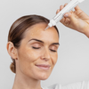 Eine Frau mit geschlossenen augen trägt das P.I.H. Sun Spot Fading Treatment auf die Stirn auf und lächelt dabei sanft.