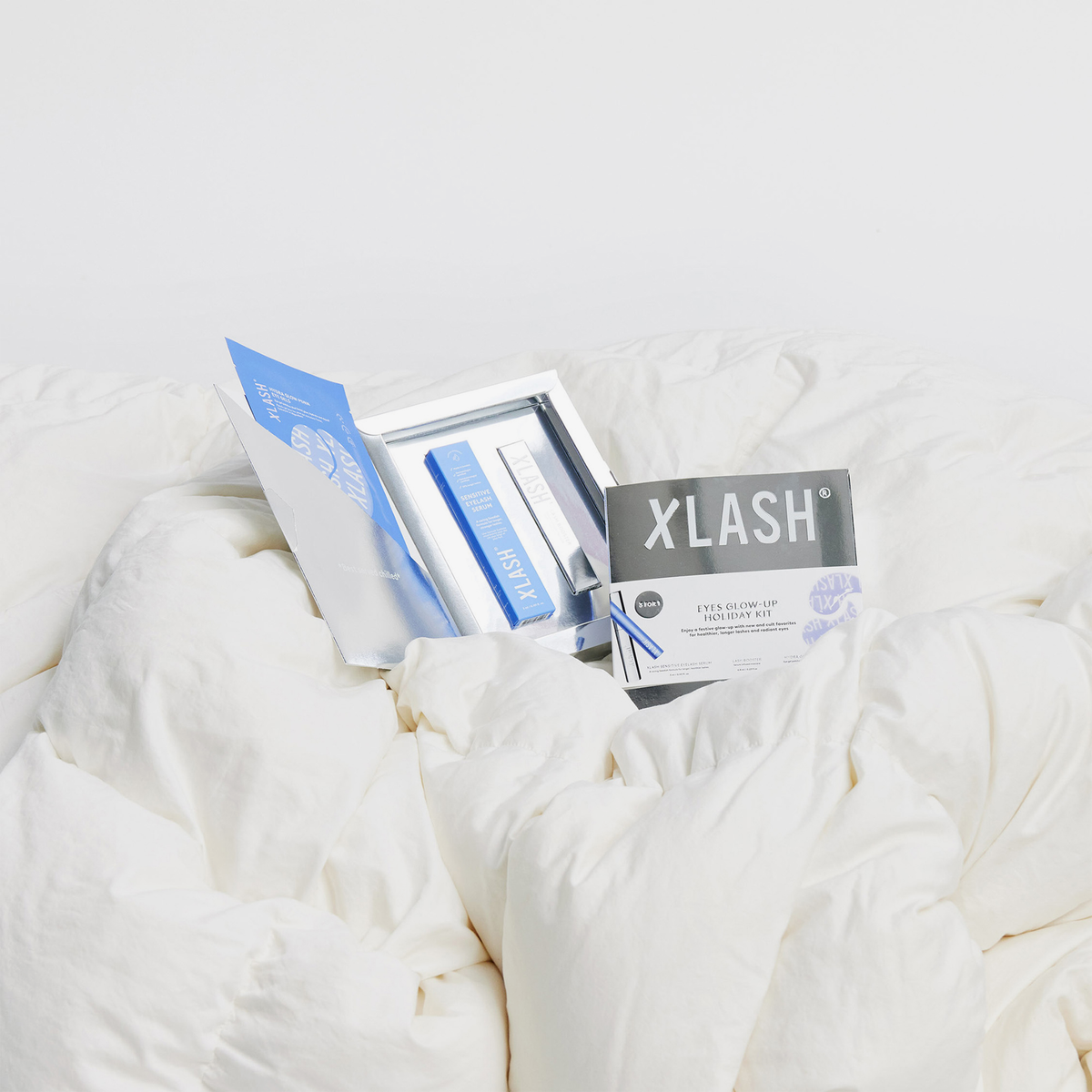 Die Verpackung des XLASH Eyes Glow-up Holiday Kit sowie das ausgepackte Set werden auf einer weichen, weißen Bettdecke gezeigt.