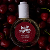 Eine Flasche Very Cherry von Hair Syrup mit Wassertropfen liegt auf dunklen Kirschen.