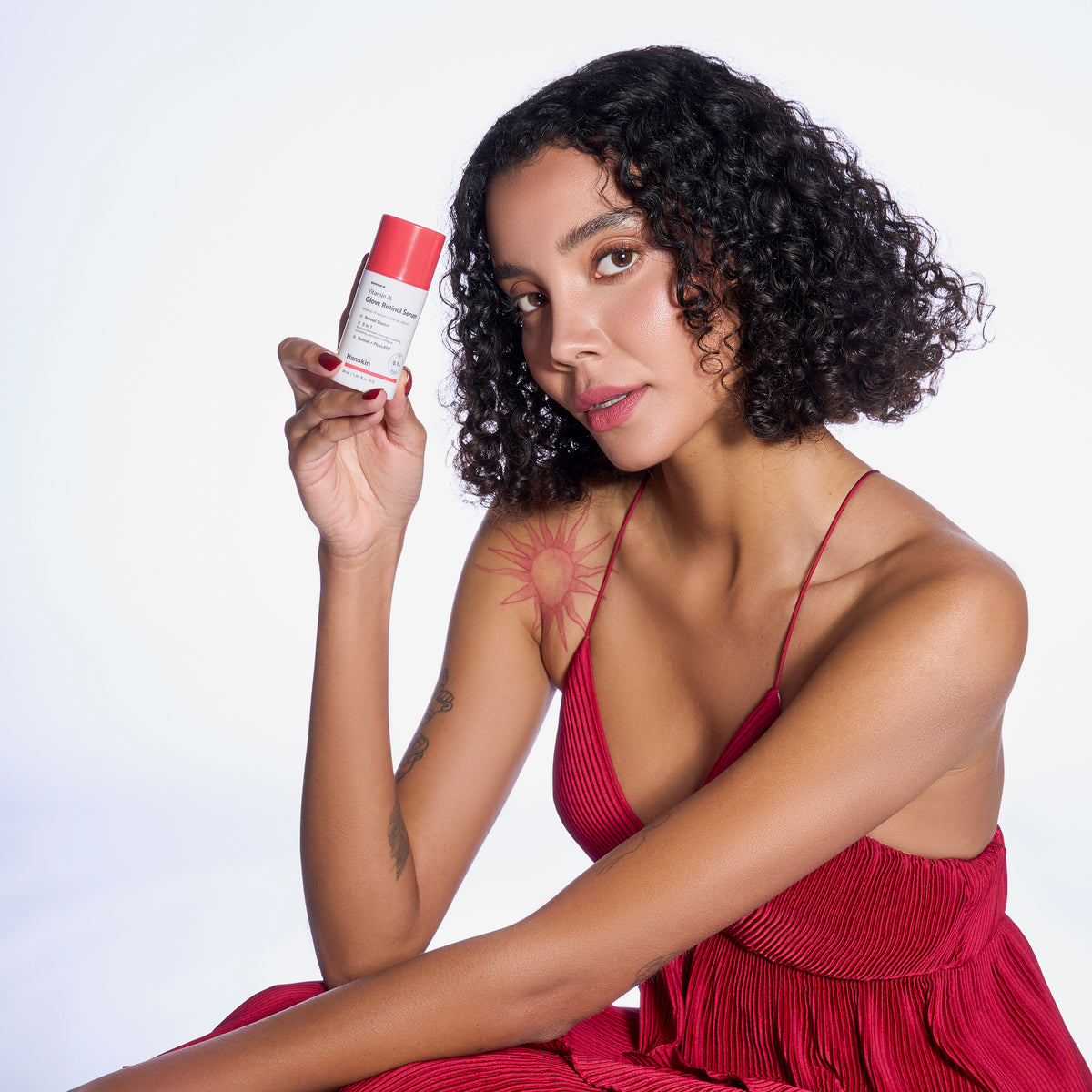 Frau in einem roten Kleid hält eine Flasche Vitamin A Glow Retinol Serum und posiert vor einem weißen Hintergrund.
