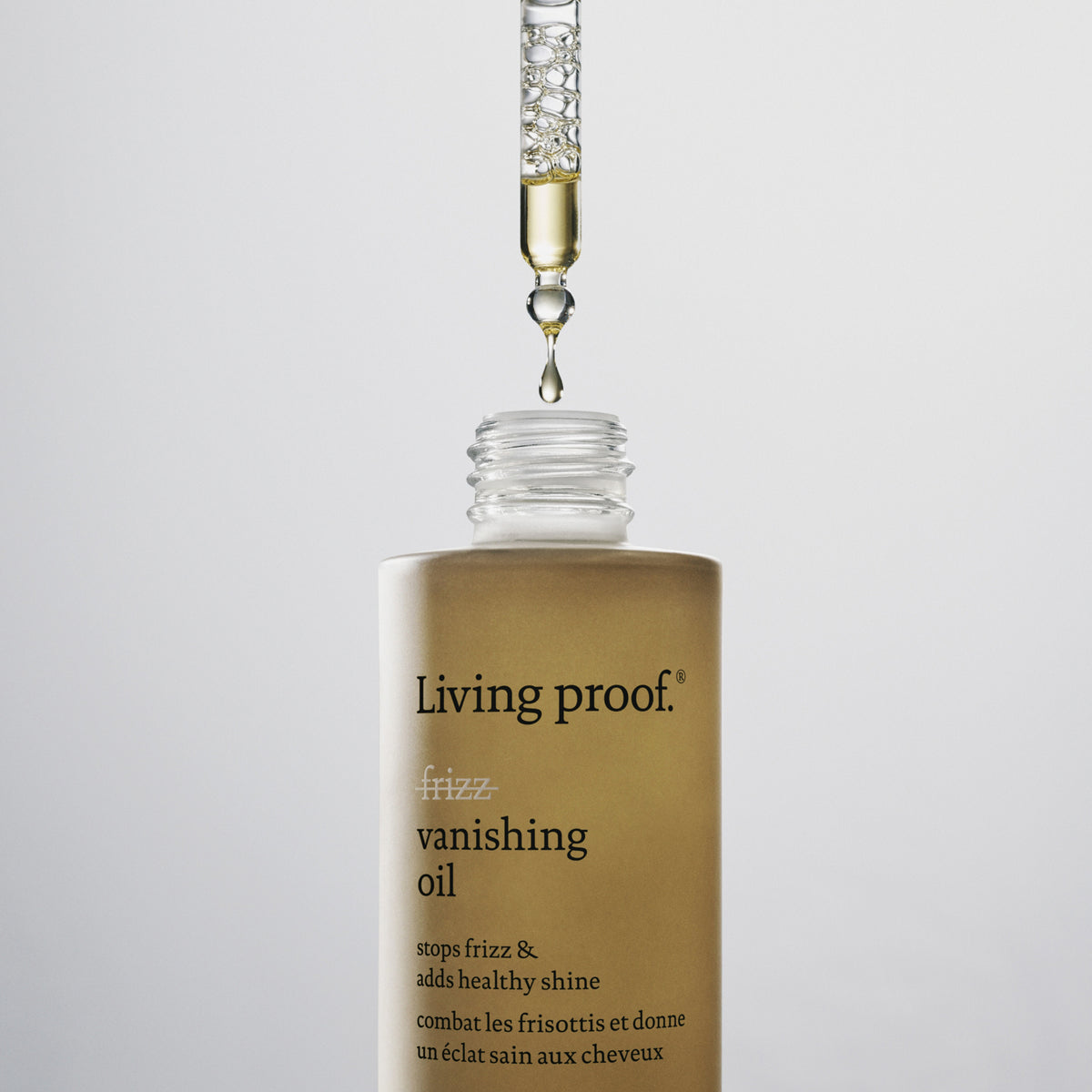 Vor einem schlichten Hintergrund gibt eine Pipette Öl in eine Flasche „Living Proof No Frizz Vanishing Oil“ ab.