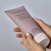 Hände halten eine Tube Living Proof Restore Repair Mask vor einem neutralen Hintergrund.