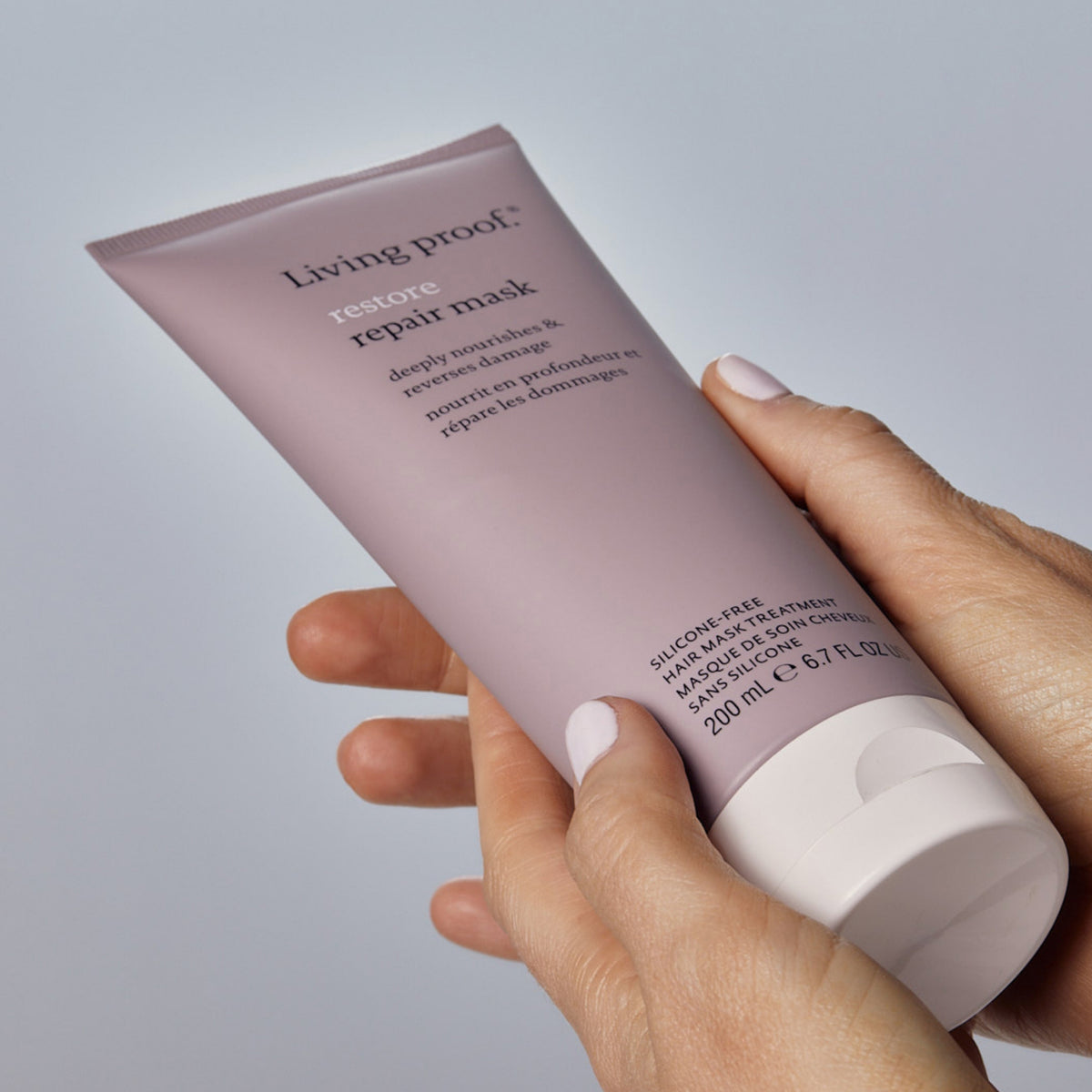 Hände halten eine Tube Living Proof Restore Repair Mask vor einem neutralen Hintergrund.
