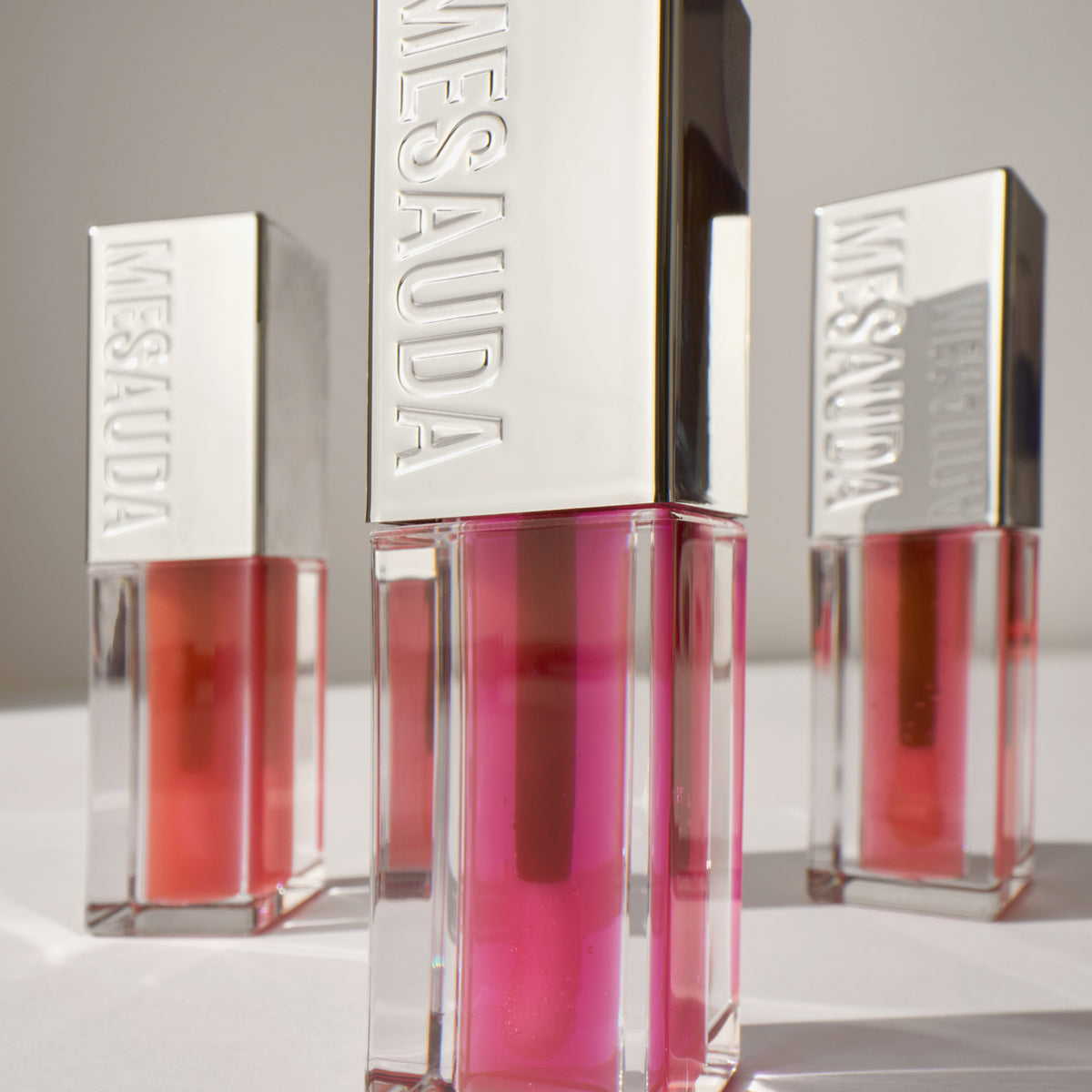Drei Flaschen Mesauda Beauty LIPOILOGY™ Lipgloss mit silbernen Kappen, in verschiedenen Tönen auf einer weißen Fläche.