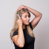 Eine Frau in einem schwarzen Shirt benutzt den Scandinavian Biolabs Scalp Activation Derma Roller, um ihr langes blondes Haar vor einem schlichten Hintergrund zu stimmulieren.