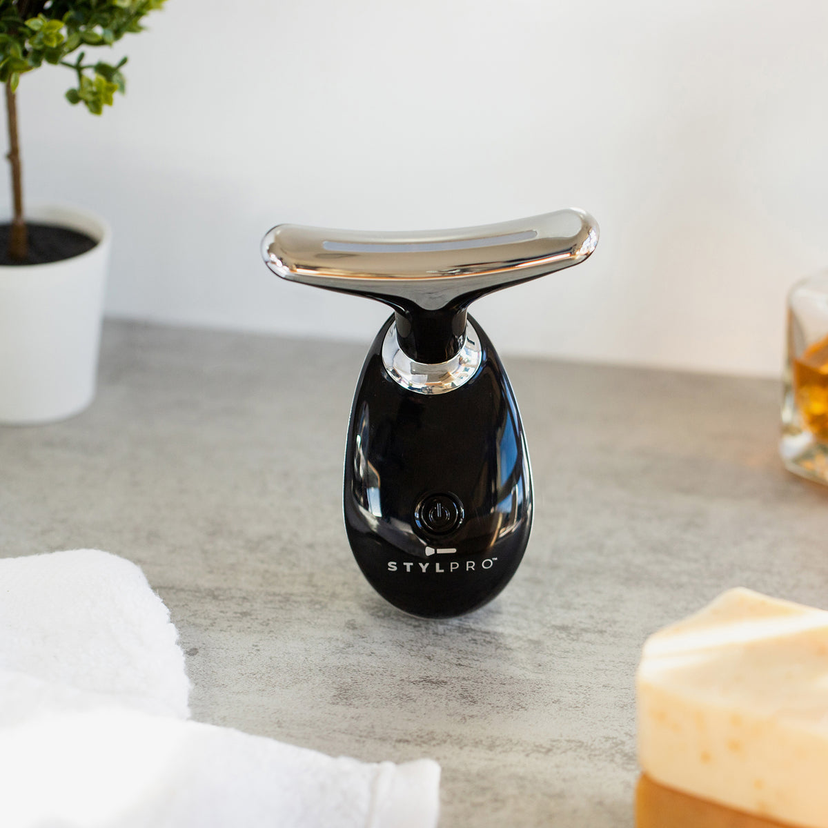 Der Fabulous Firmer Neck & Face Smoother in Schwarz und Silber steht auf einer Arbeitsplatte neben einer Topfpflanze und Seife.