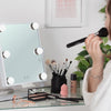 Eine Frau, die sich an ihrem Schminktisch schminkt, zeigt den Glam & Groove Hollywood Bluetooth Mirror, umgeben von Kosmetika und einer grünen Pflanze.