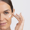 Eine Frau trägt vor einem weißen Hintergrund mit zwei Fingern eine Barrier Restoring Hydrating Cream auf ihre Wange auf.
