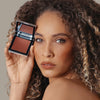 Eine Frau hält den geöffneten Skin Mate Compact von Mesauda Beauty in der Nähe ihres Gesichts und blickt in Kamera.