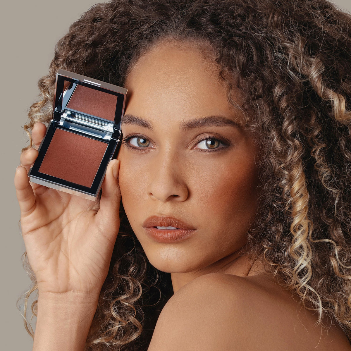 Eine Frau hält den geöffneten Skin Mate Compact von Mesauda Beauty in der Nähe ihres Gesichts und blickt in Kamera.