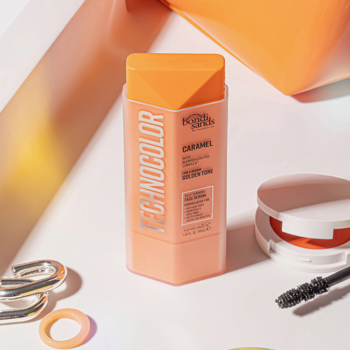 Eine Flasche Technocolor Self Tanning Face Serum Caramel auf einer weißen Fläche, umgeben von Make-up-Artikeln.