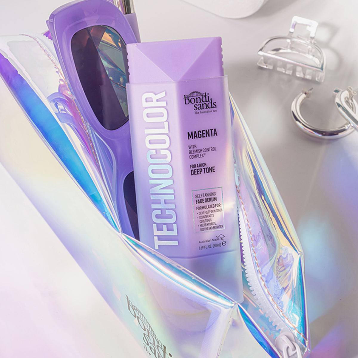 Lila Flasche Technocolor Self Tanning Face Serum in einer Kosmetiktasche mit Sonnenbrille; umgeben von weiteren Accessoires.