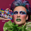 Eine Person mit hellem Teint und buntem Haar zeigt kräftiges, farbenfrohes Make-up hält die Love is Love-Palette.