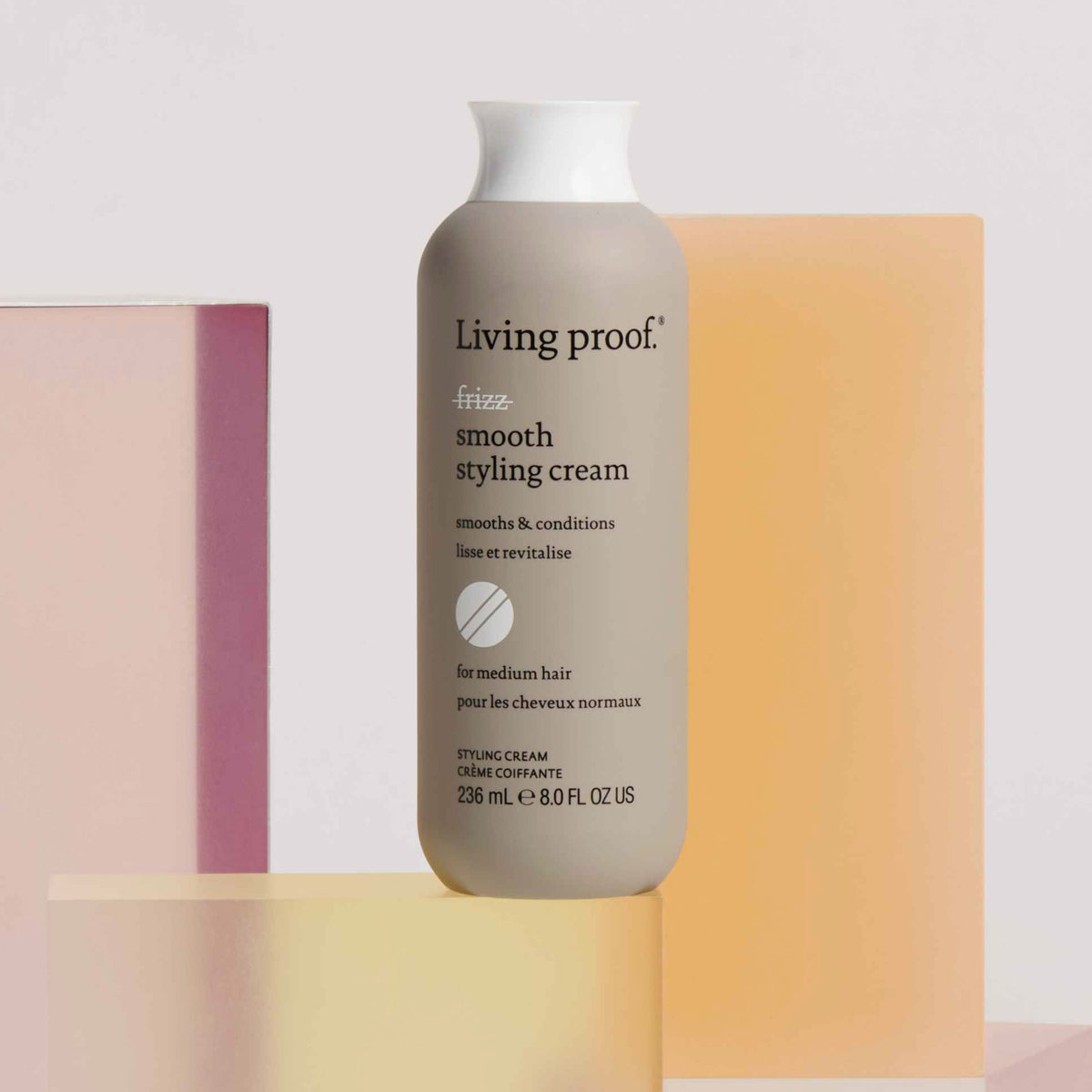 Eine Flasche Living Proof No Frizz Smooth Styling Cream wird vor pastellfarbenen geometrischen Formen präsentiert.