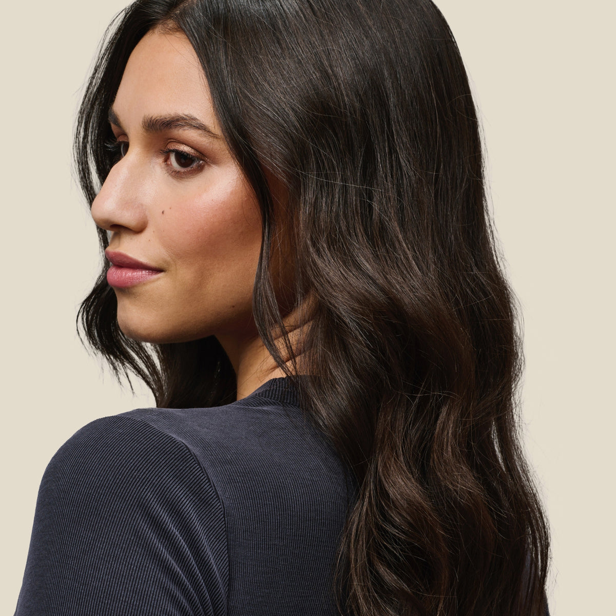 Eine Frau mit langem, welligem, dunklem Haar, gestylt mit Living Proof No Frizz Vanishing Oil, vor einem hellen Hintergrund.