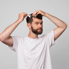 Ein bärtiger Mann in einem weißen T-Shirt benutzt den Scandinavian Biolabs Scalp Activation Derma Roller und bürstet sein Haar vor einem schlichten hellgrauen Hintergrund.