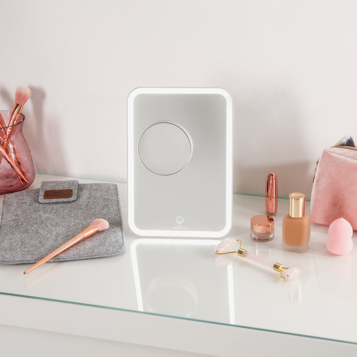 Schminktisch aus Glas auf dem der Glow & Go-Reisespiegel, Make-up-Pinsel, Foundation, Lippenstift und einem rosa Beutel präsentiert sind.