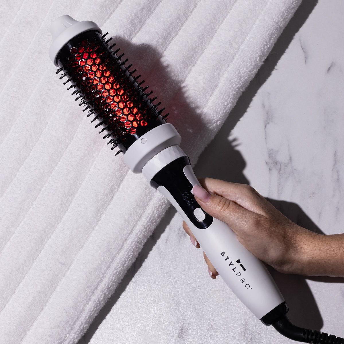 Eine Hand hält die STYLPRO Nahinfrarot-Haarbürste mit weißem Griff, die neben einem weißen Handtuch abgebildet ist.