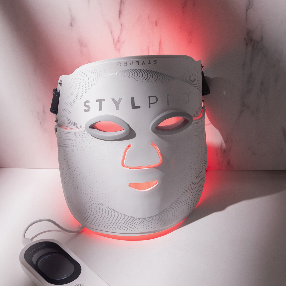 Die STYLPRO Wavelength Pro 5 in 1 LED-Maske leuchtet von hinten rot, während sie auf einer Marmoroberfläche liegt und die Fernbedienung in der Nähe ist.