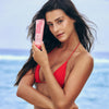 Frau in einem roten Bikini, die eine rosa Flasche Rapid Warming Balm hält, im Hintergrund das Meer.