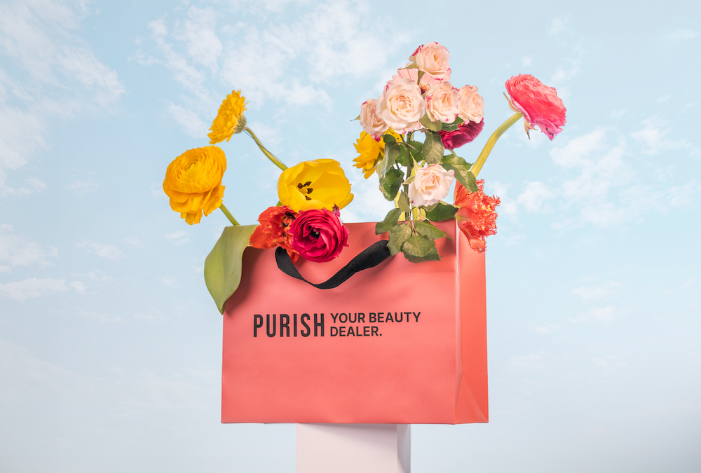 Blumen in leuchtenden Farben stecken in einer rosa Einkaufstasche mit der Aufschrift ‚PURISH YOUR BEAUTY DEALER‘ vor Himmel-Hintergrund.