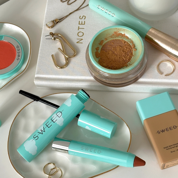 Make-up-Artikel in türkisfarbenen Verpackungen, Goldschmuck, ein NOTES-Notizbuch und Puder auf einem weißen Tisch.