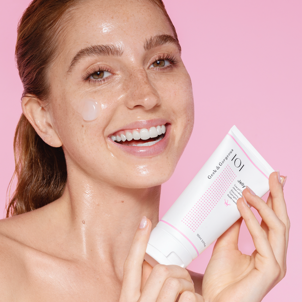 Eine lächelnde Frau trägt Hautpflegecreme auf ihre Wange auf, während sie eine weiß-rosa Tube vor einem rosa Hintergrund in der Hand hält.