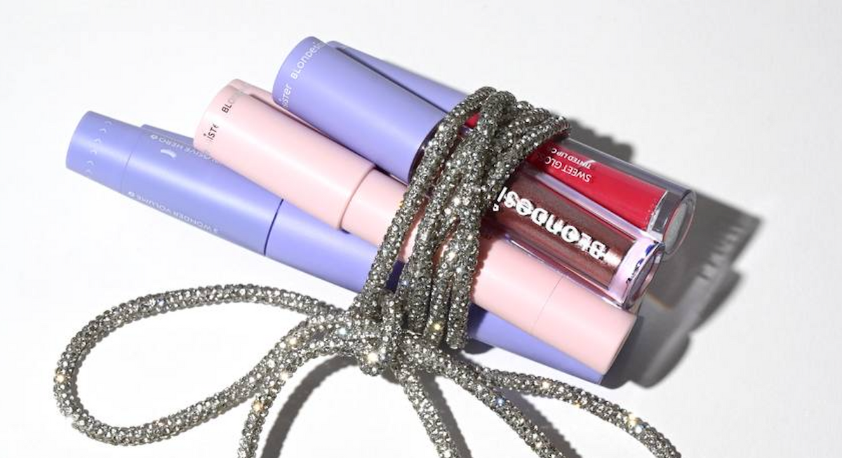 Drei pastellfarbene Make-up-Tuben und ein roter Lipgloss sind mit einer glitzernden silbernen Schnur auf einem weißen Untergrund umwickelt.
