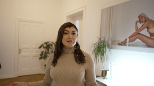 Büsra in beigefarbenem Rollkragenpullover steht in modernem Raum mit Pflanzen und großem Wandbild im Hintergrund.