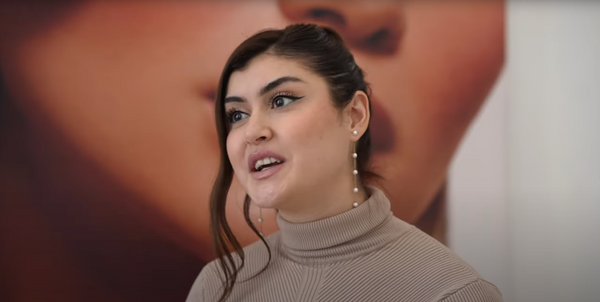 Büsra trägt lange Perlenohrringe, einen beigefarbenen Rollkragenpullover und spricht, im Hintergrund ein unscharfes Porträt.