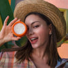 Eine Frau mit Strohhut lächelt in die Kamera und hält einen orangefarbenen Behälter mit Pore Cleansing Balm AHA hoch.