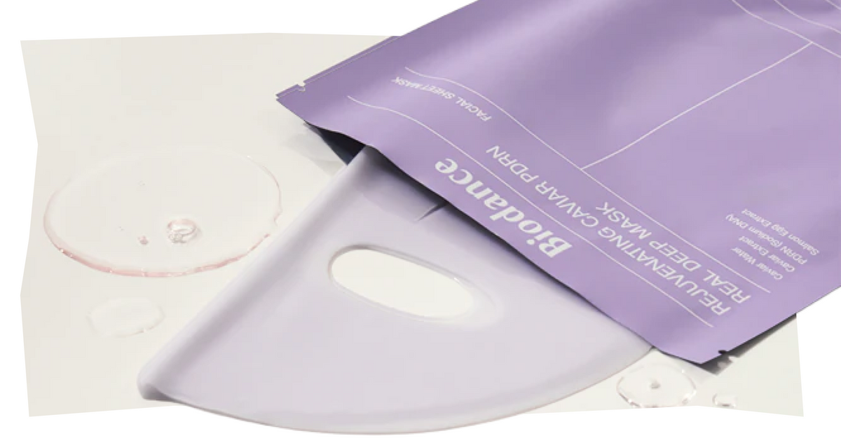 Eine teilweise geöffnete Verpackung einer violetten Gesichtsmaske, daneben auf einer weißen Fläche Tropfen einer klaren Flüssigkeit.
